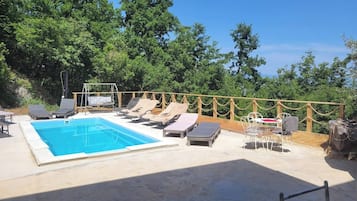 A swimming pool with lounge chairs and a table, surrounded by a wooden deck and trees.