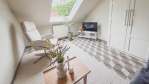 Ein modernes Schlafzimmer im Loftsstil mit schrägverdachter Decke, einem großen Fenster, einem weißen Kleiderschrank, einem Fernseher auf einem Schrank und einem kleinen Tisch mit Vase und Kerze.