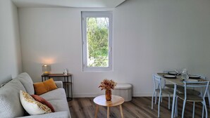 Uma sala de estar moderna com um sofá, uma mesa redonda pequena, uma mesa de jantar com cadeiras e uma janela com vista para o verde.
