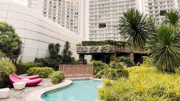 A modern building with a pool, surrounded by greenery and pink lounge chairs. - Sky Suite-City Sunset & River views (Mandaluyong City)