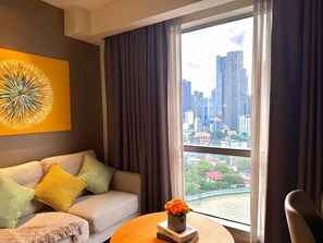 A modern hotel room with a large window, a sofa, a table with a vase of flowers, and a cityscape view. - Sky Suite-City Sunset & River views (Mandaluyong City)