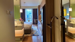 A hotel room with a bed, bedside tables, a desk, a chair, a bathroom with a mirror, and a view of the cityscape. - Sky Suite-City Sunset & River views (Mandaluyong City)