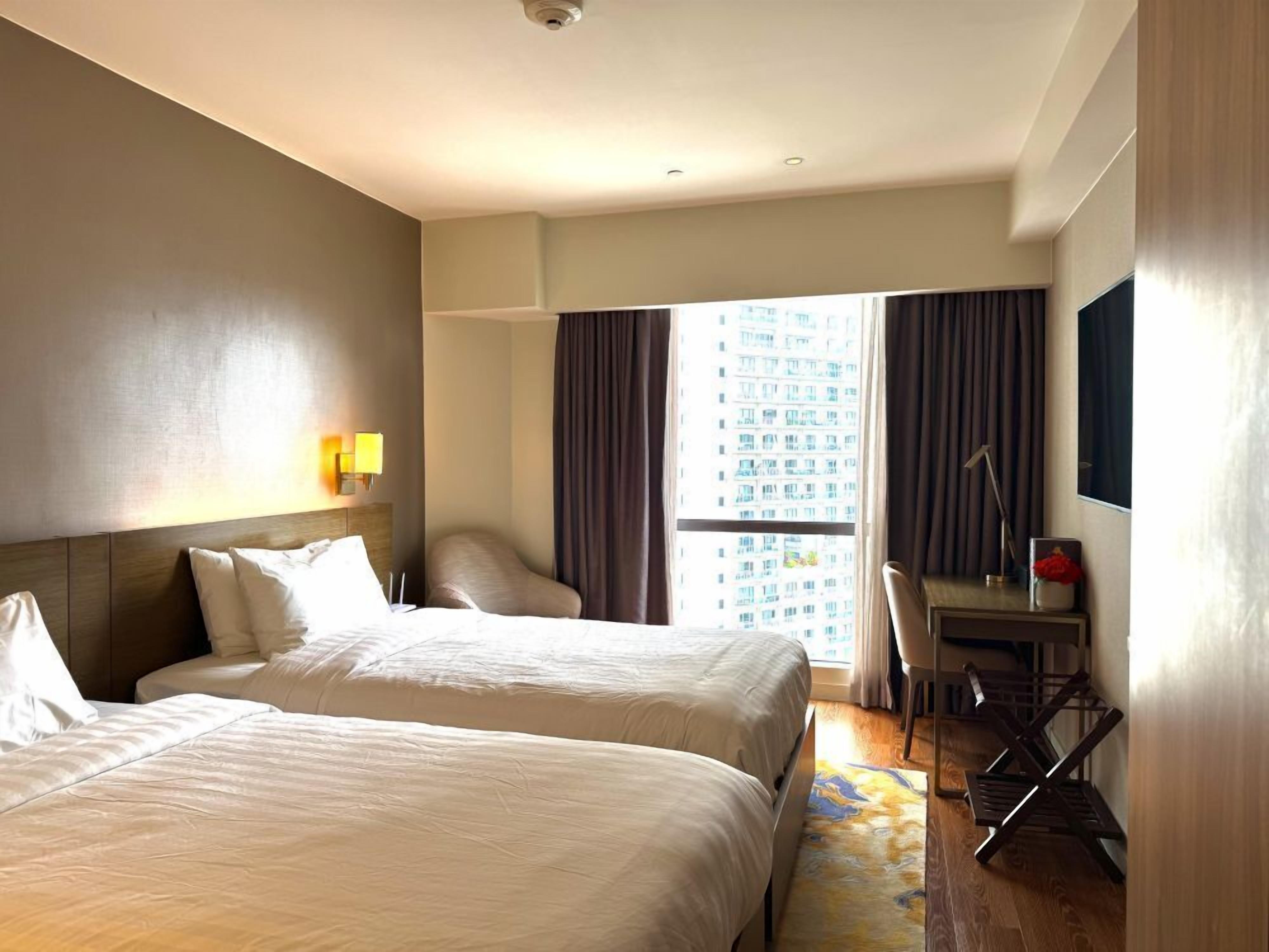 A hotel room with two beds, a desk, a chair, a lamp, and a large window with curtains.