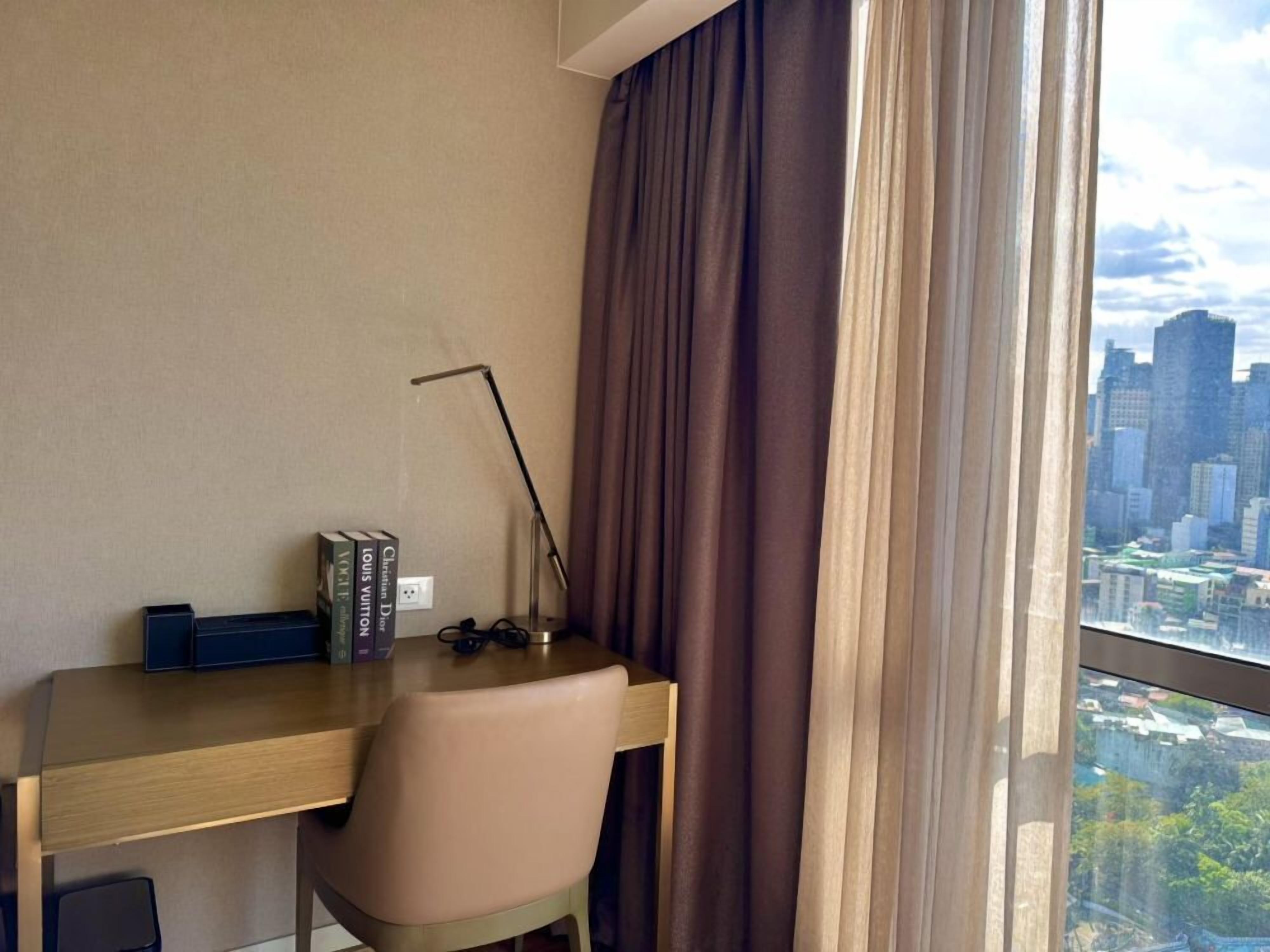 Habitación de hotel con escritorio, silla, libros y vistas a un paisaje urbano a través de una ventana.