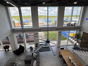 A modern interior with large windows, a central fan, a dining area, and a view of a river and outdoor seating.