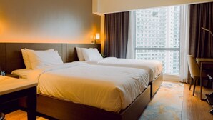 Une chambre d’hôtel avec deux lits, un bureau, une chaise et une grande fenêtre donnant sur des immeubles de grande hauteur.