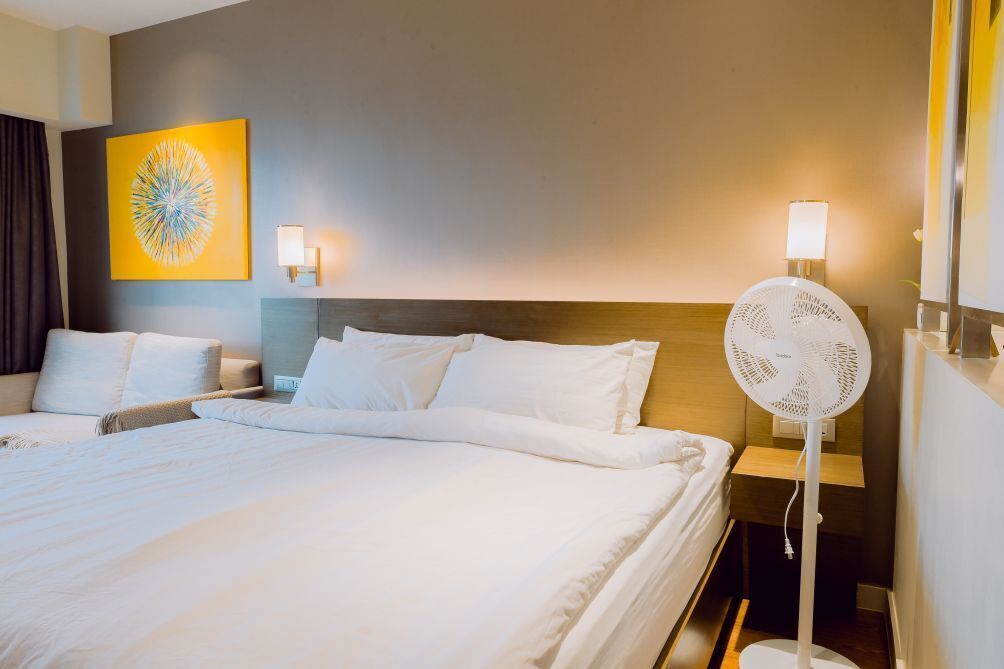 Une chambre d’hôtel avec deux lits, un ventilateur sur pied, une lampe fixée au mur et une œuvre d’art abstraite.