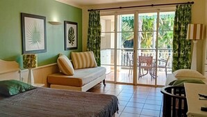 Een kamer met een bed, een bank, een eettafeltje en een stoel. Twee ingelijste foto’s hangen aan de muur, en er is een balkon met uitzicht op groen.