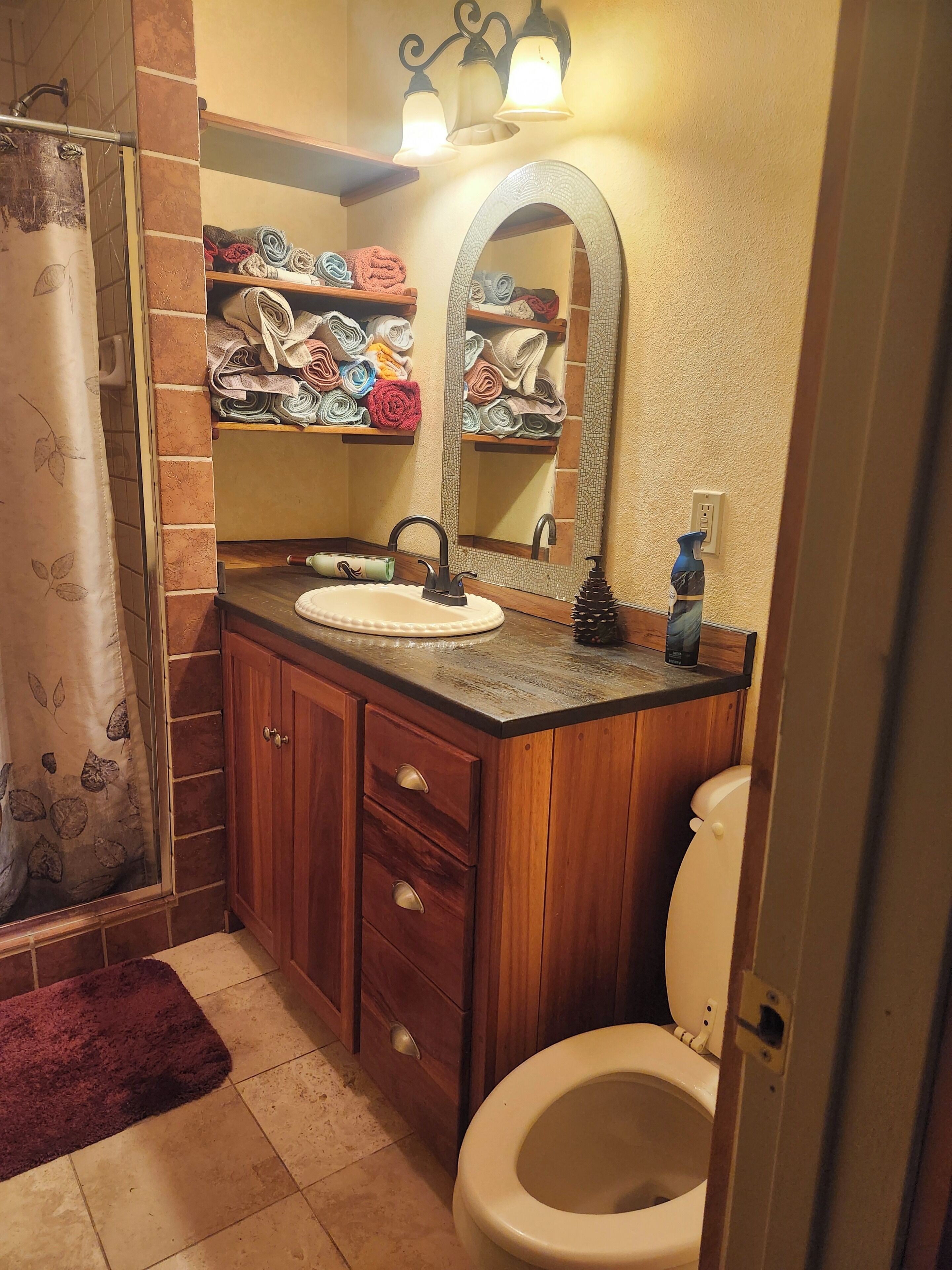 Un baño con ducha, lavabo con espejo, inodoro y estante con toallas.
