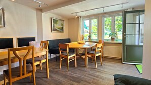 A room with a dining table, chairs, a sofa, a painting on the wall, and a window with a view of greenery.