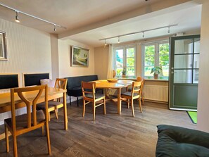 Ein Zimmer mit Esstisch, Stühlen, einer Couch, einem Bild an der Wand und einem Fenster mit Blick auf Grünflächen.