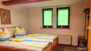 A bedroom with a wooden bed, two windows with green blinds, a radiator, a small table, and a trash bin.