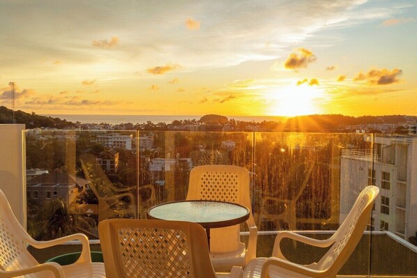 A balcony with a table and chairs, overlooking a cityscape at sunset. - Famous Kata Ocean View 2BR Seaview Residence c129 (Tambon karon)
