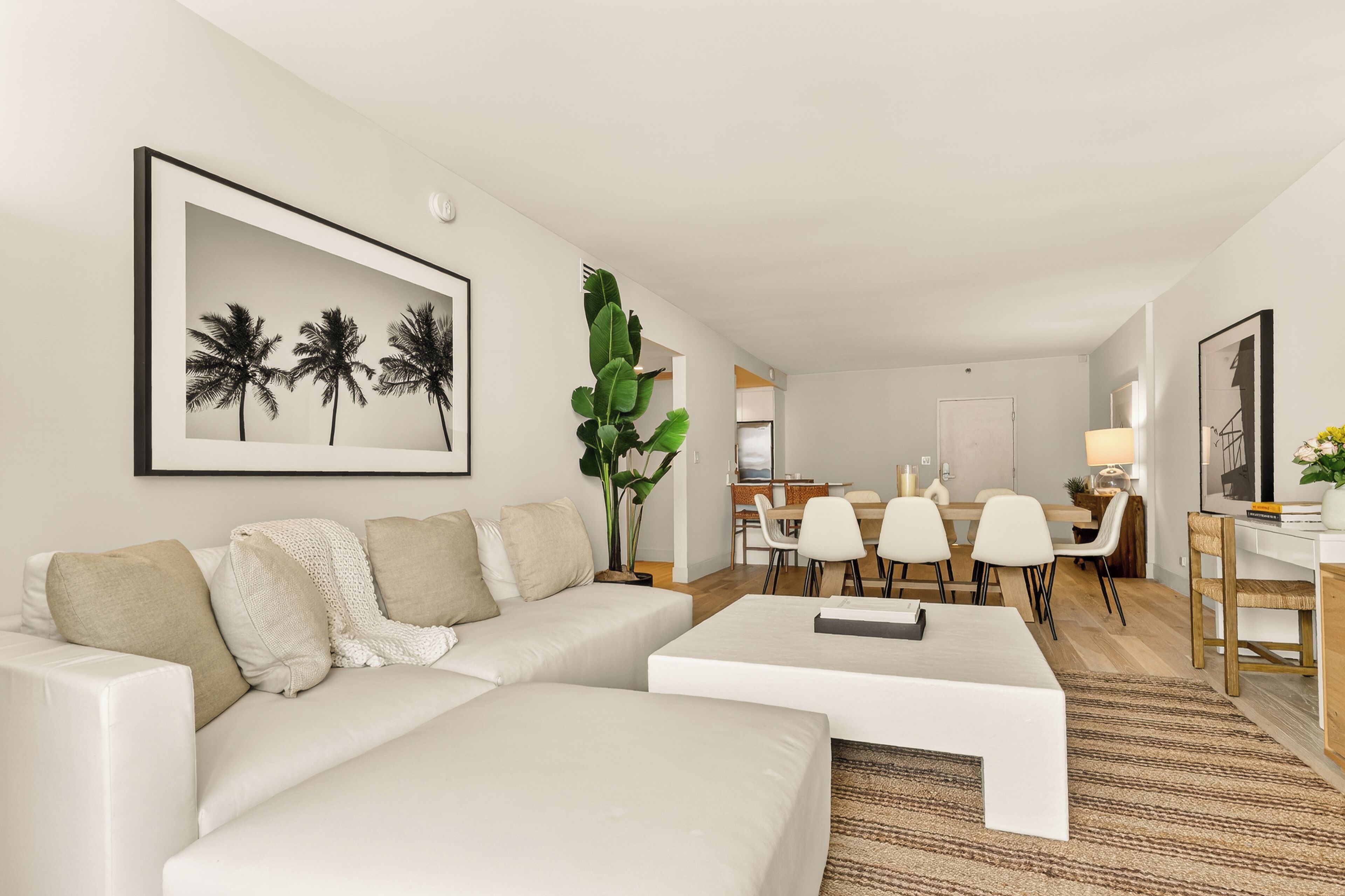 A modern living room with a sofa, coffee table, dining area, and a large framed picture of palm trees.
