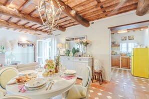 Ein Essbereich mit einem runden Tisch, Stühlen und einer Pendelleuchte. Im Hintergrund ist eine Küche mit Holzschränken und einem gelben Kühlschrank zu sehen.