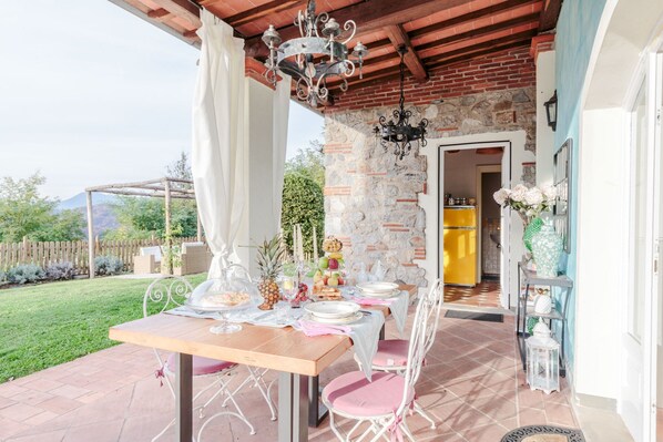 Ein Essbereich im Freien mit einem Tisch für vier Personen, umgeben von weißen Stühlen, einer Steinmauer und einer Holzpergola.