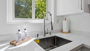 Una cocina moderna con un lavamanos de acero inoxidable, una ventana con vistas a árboles y dos botellas con las etiquetas 'DISH' y 'HAIR'.