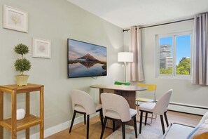 Ein modernes Zimmer mit einem runden Tisch, zwei Stühlen, einem Fernseher, einer Lampe, einem Fenster mit Vorhängen und gerahmten Bildern an der Wand.