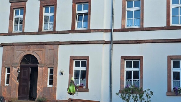 Een gebouw met twee verdiepingen, een symmetrische gevel, een centrale ingang met een deur en een kleine tuin met een bankje en plantenpotten.
