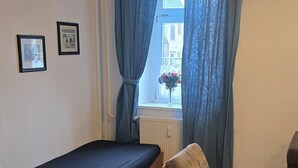 Een kamer met een bed, een stoel, een raam met gordijnen en twee afbeeldingen in lijsten aan de muur.