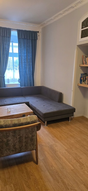 A room with a sectional sofa, a wooden coffee table, a patterned armchair, a bookshelf, and a window with curtains.