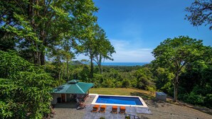 Piscina com vista para o oceano, cercada por vegetação exuberante e um espaço de estar coberto.