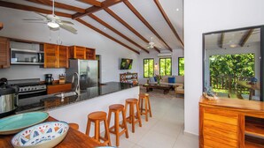 Cozinha moderna com armários de madeira, uma ilha grande com banquetas, geladeira e área de refeições com vista para o verde.