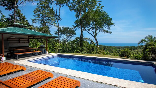 Piscina com espreguiçadeiras de madeira, área de estar coberta e vista para o oceano.