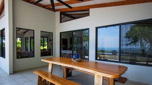 Uma área de jantar moderna com mesa e bancos de madeira, grandes janelas com vista para o exterior e teto alto com vigas aparentes.