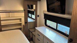 A compact RV interior with a bed, a TV mounted on a wall, and a window with an exit sign. - Unique Country Getaway Luxury Peaceful & Spacious (Cleveland)