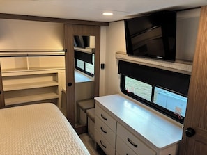 Ein kompakter Wohnraum in einem Wohnmobil mit einem Bett, einem an der Wand montierten Fernseher und einem Fenster mit einem Fluchtzeichen.