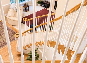 Una escalera con balaustres blancos y peldaños de madera que conduce a un salón con un sofá, una alfombra roja y una planta.