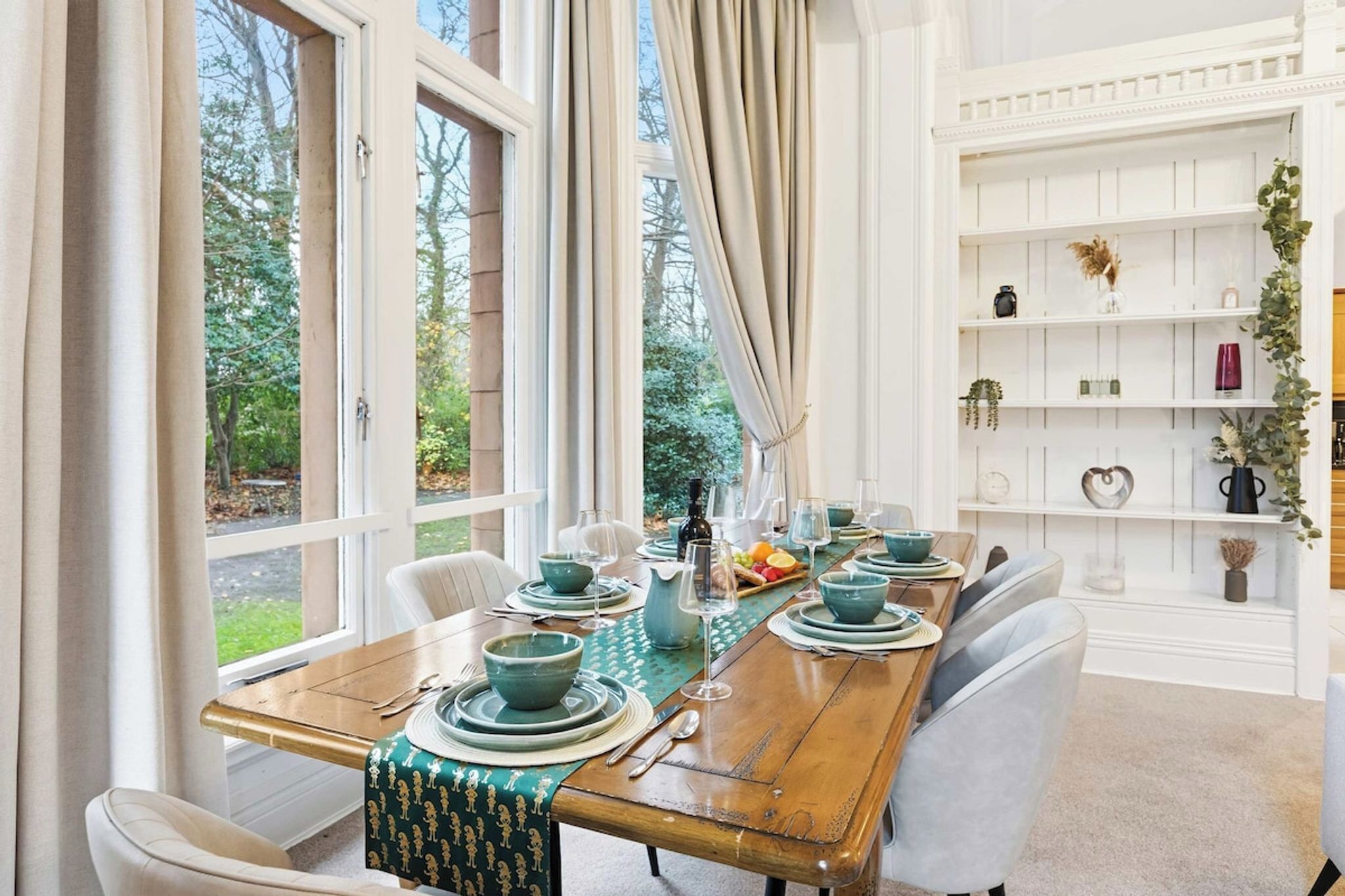 A dining room with a wooden table set for four, surrounded by white chairs, with a view of greenery outside.