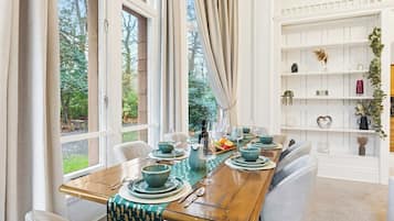 A dining room with a wooden table set for four, surrounded by white chairs, with a view of greenery outside.