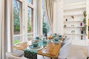 Une salle à manger avec une table en bois dressée pour quatre personnes, entourée de chaises blanches, et offrant une vue sur la verdure à l’extérieur.