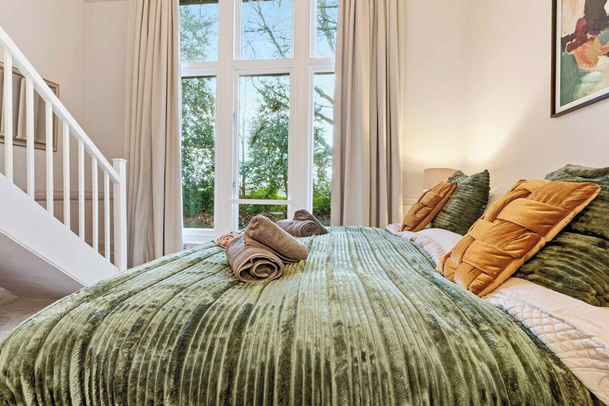 A bedroom with a large window, a bed with green bedding, and a staircase.