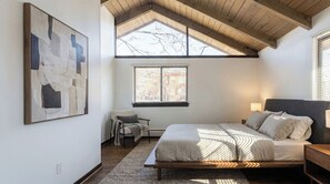Ein Schlafzimmer mit schrägem Dach, einem Bett, einem Nachttisch, einem Sessel, einem Fenster mit Jalousien und einem großen abstrakten Gemälde an der Wand.