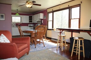 A cozy living room with a red sofa, wooden chairs, a dining table, and a kitchen area with white cabinets and a large window. - Lovely 2-bedroom cottage in Fletcher perfect for a relaxing getaway (Fletcher)