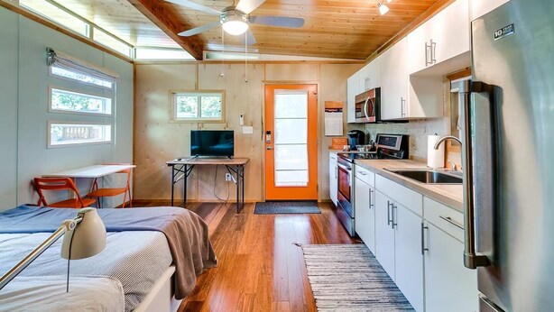 Ein kleines Zimmer mit Bett, Schreibtisch, Stuhl, Fernseher, Küchenzeile und Deckenventilator.