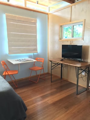 Ein kleiner Raum mit einem Holztisch, zwei orangefarbenen Stühlen, einem Fernseher und einem Fenster mit Jalousien.