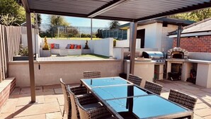 An outdoor patio with a glass-top table and wicker chairs, a brick oven, and a barbecue grill. - Large detached home for families and group stays (Hoyland)