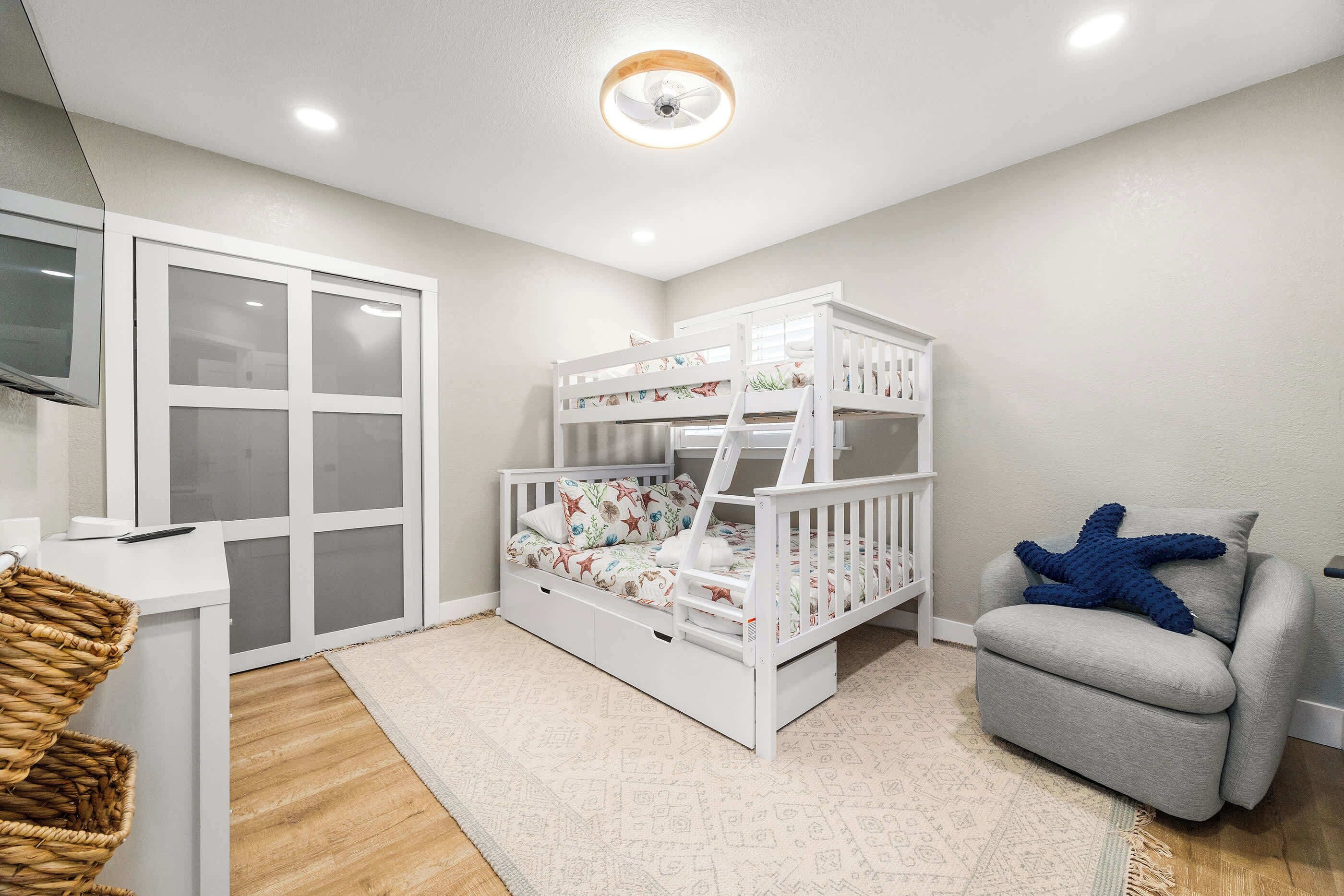 Un lit superposé pour enfant avec un coussin en forme d’étoile de mer, un fauteuil gris, un bureau blanc et un panier en osier.