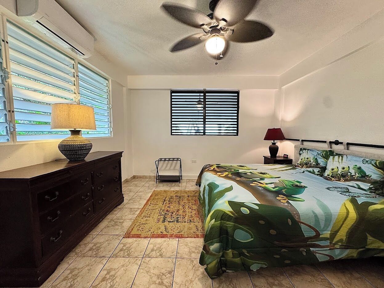Un dormitorio con cama, ventilador de techo, ventana con persianas, cómoda y mesita de noche con lámpara.