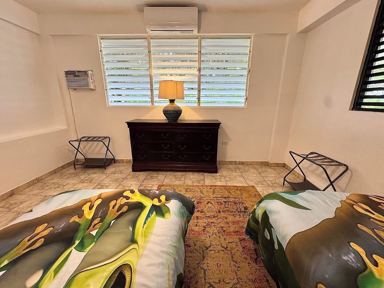 Una habitación con dos camas, un aparador, una lámpara y una ventana con persianas.