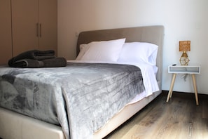 Un lit bien fait, recouvert d’une couverture grise, une pile de serviettes pliées et une lampe de chevet posée sur une table de nuit en bois.