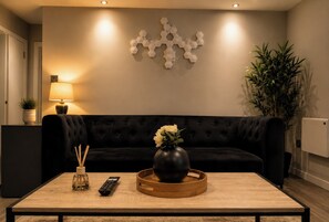 A modern living room with a black sofa, a wooden coffee table, a vase with flowers, a remote control, and a wall-mounted decorative piece. - Canal-Side Apartment with Private Terrace - Castlefield - Business & Long Stays (Greater Manchester)