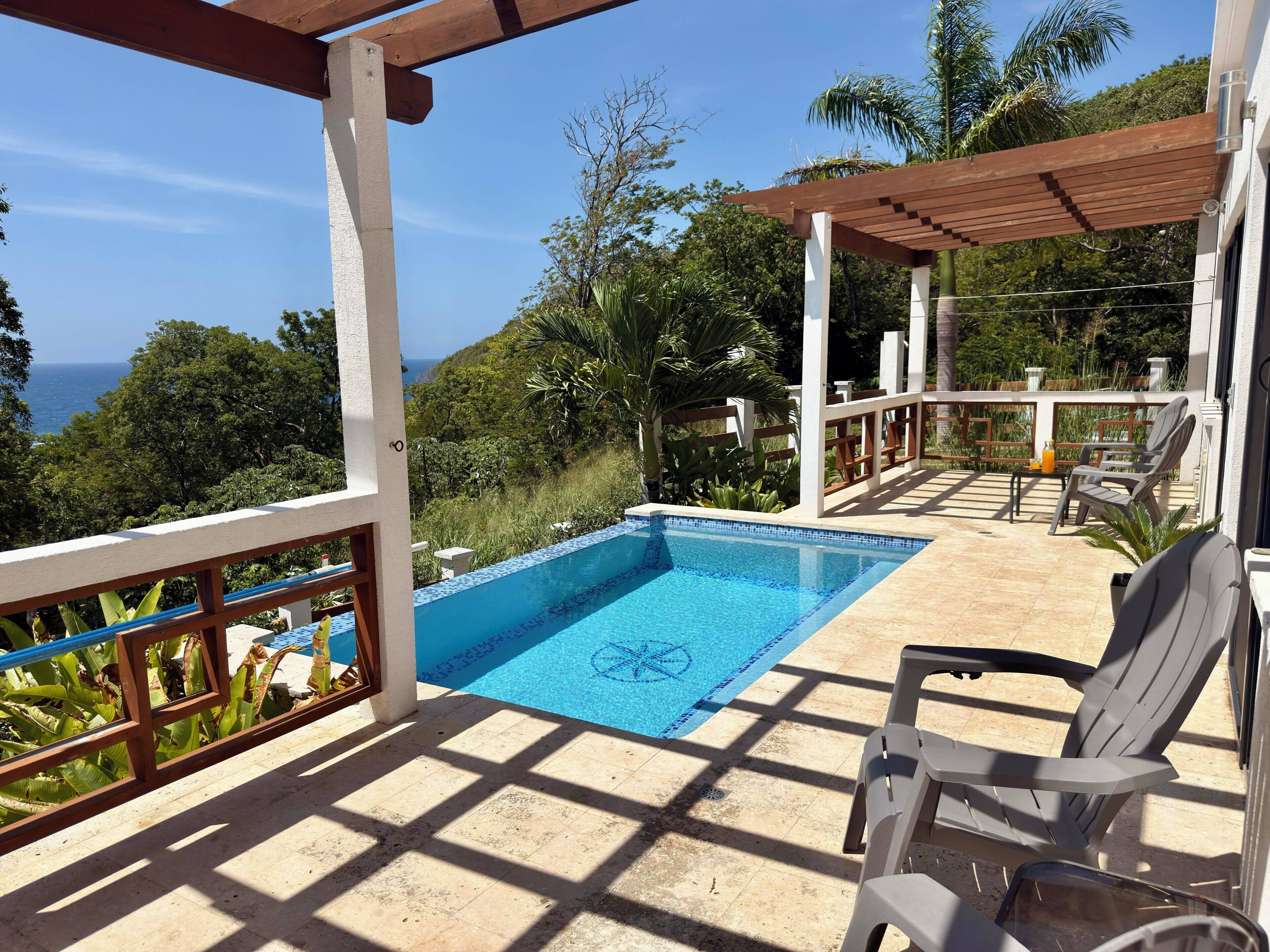 Una piscina con vistas al océano, rodeada de vegetación y una pérgola.