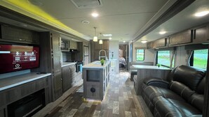 A compact RV interior with a kitchenette, a central island, a seating area, and a TV mounted on the left wall. - EastToWest Glamping camper - Site #15 (Klamath)