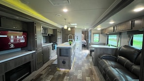 Interior compacto de autocaravana con kitchenette, una isla central, zona de estar y televisor montado en la pared izquierda.