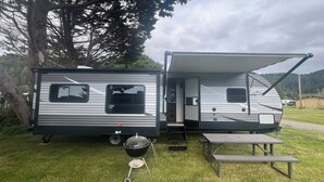 A travel trailer with a covered awning, a small enclosed kitchen area, and a picnic table. - EastToWest Glamping camper - Site #15 (Klamath)